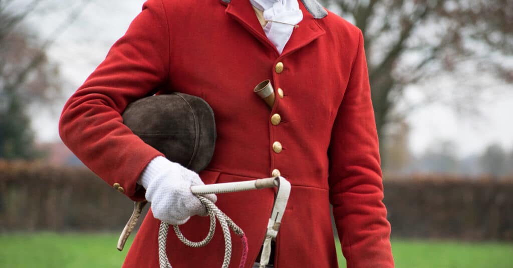 Person wearing red foxhunting jacket. We can see just the jacket and arms. They are holding a riding helmet and hunting whip.